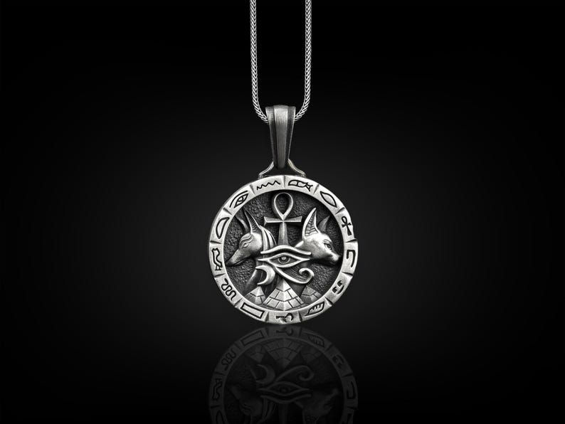 Pu&ograve; includere: Collana con ciondolo in argento di design egizio. Il ciondolo &egrave; un cerchio con un ankh, l'occhio di Horus e due teste di sciacallo. Il cerchio &egrave; circondato da geroglifici.