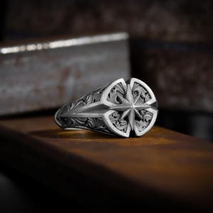 May include: A silver ring with an intricate design featuring a compass rose in the center. The ring is set against a rustic background.