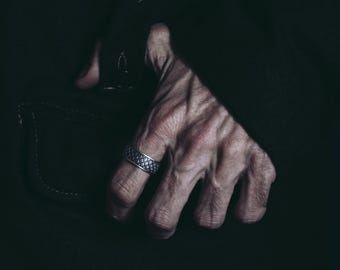 May include: A close-up of a hand wearing a silver ring with a textured design. The hand is against a dark background.