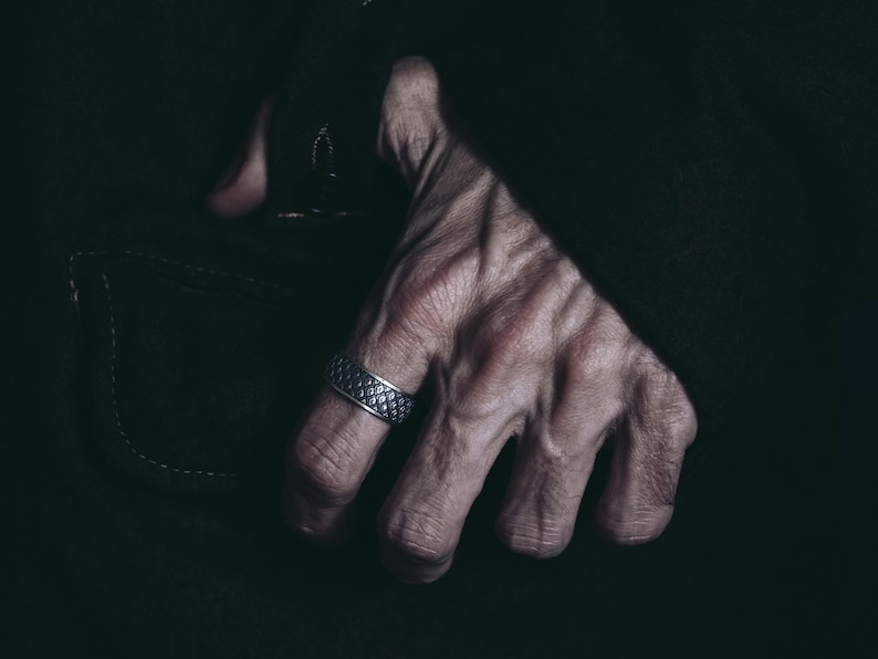 K&ouml;nnte beinhalten: Nahaufnahme einer Hand, die einen silbernen Ring mit einem strukturierten Design tr&auml;gt. Die Hand befindet sich vor einem dunklen Hintergrund.