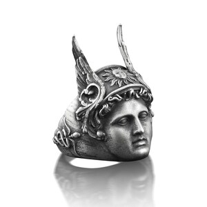 May include: A silver ring with a detailed design of a woman's face wearing a winged helmet with a sun symbol. The ring is a statement piece with intricate details.