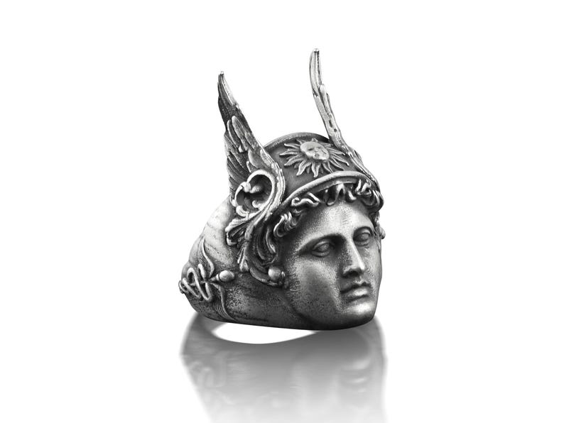 May include: A silver ring with a detailed design of a woman's face wearing a winged helmet with a sun symbol. The ring is a statement piece with intricate details.