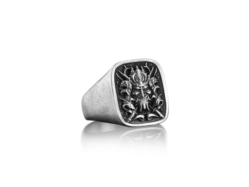 May include: A silver ring with a square face featuring a detailed, black and white engraving of a samurai warrior's mask.