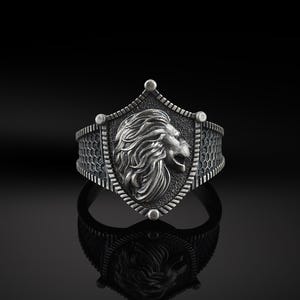 May include: A silver ring featuring a lion head design on a shield. The ring has a textured band.