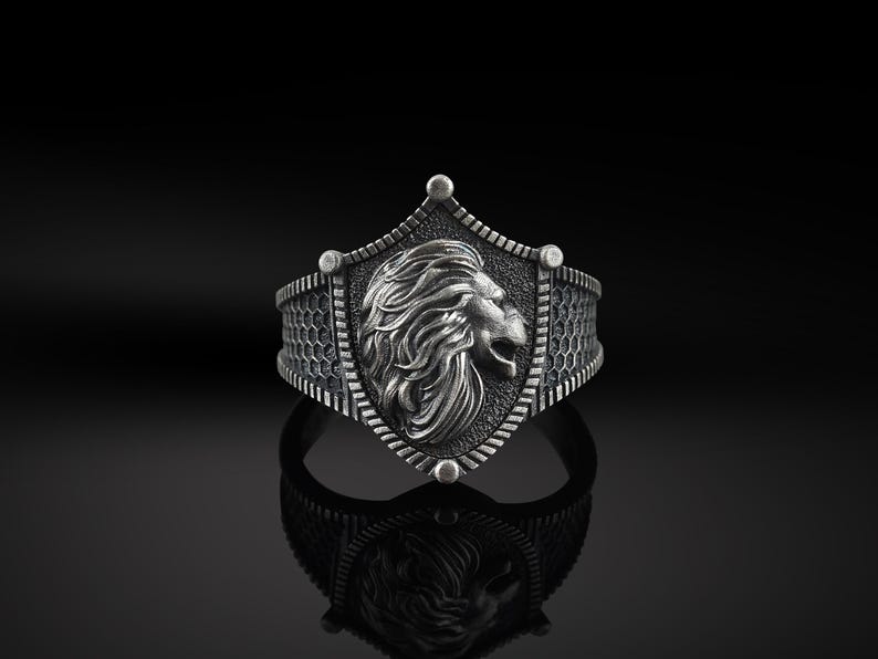 May include: A silver ring featuring a lion head design on a shield. The ring has a textured band.