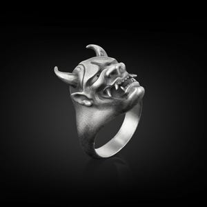 May include: A silver ring with a detailed, three-dimensional design of a demon's face. The ring is made of metal and has a textured finish.