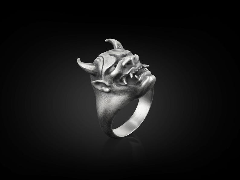 May include: A silver ring with a detailed, three-dimensional design of a demon's face. The ring is made of metal and has a textured finish.