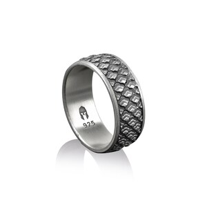 K&ouml;nnte beinhalten: Ein silberner Ring mit einem strukturierten, gemusterten Band. Das Band ist dunkelgrau und hat ein erhabenes Design. Die Innenseite des Rings ist mit "925" und einem kleinen Helm-Symbol gepr&auml;gt.