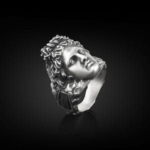 Peut inclure: Une bague en argent avec une sculpture d&eacute;taill&eacute;e d'un visage humain. Le visage regarde vers le haut et vers la droite. La bague est en m&eacute;tal et a une finition lisse.