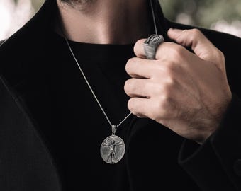 May include: A silver chain necklace with a pendant featuring a detailed depiction of the Vitruvian Man. The pendant is round and hangs from a silver chain. The person is wearing a black shirt.