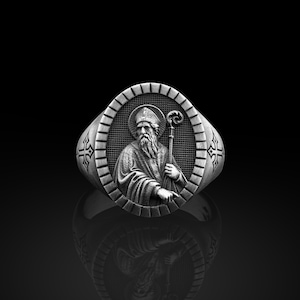 Puede incluir: Un anillo de plata con un diseño detallado que presenta una figura religiosa sosteniendo un bastón. El anillo tiene una cruz a cada lado.