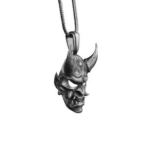 May include: A silver pendant necklace featuring a detailed, stylized oni mask with horns. The mask is a traditional Japanese demon mask.