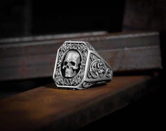 Könnte beinhalten: Ein silberner Ring mit einem detaillierten Totenkopfdesign in der Mitte. Der Ring ist in einem quadratischen Rahmen mit filigranen Blumenmustern gefasst.