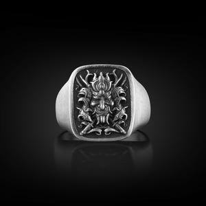 May include: A silver ring with a square face featuring a detailed design of a samurai warrior's mask with swords and flames.