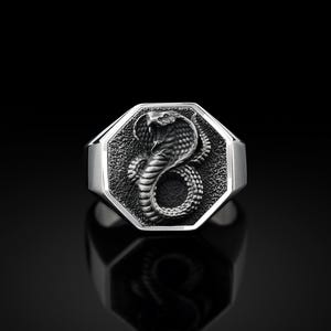 Könnte beinhalten: Ein silberner Ring mit einer achteckigen Oberfläche, die ein detailliertes, dreidimensionales Design einer Kobra-Schlange zeigt.