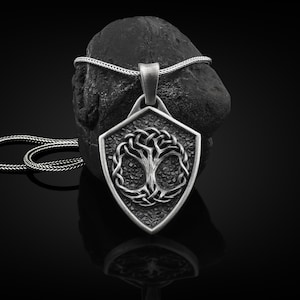 May include: A silver pendant necklace with a shield-shaped design featuring a Celtic knot tree of life. The pendant is on a black background.