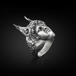May include: A silver ring with a detailed sculpted face of a winged figure wearing a helmet. The ring is set against a black background.