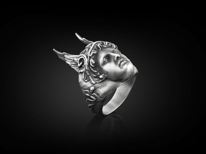 May include: A silver ring with a detailed sculpted face of a winged figure wearing a helmet. The ring is set against a black background.