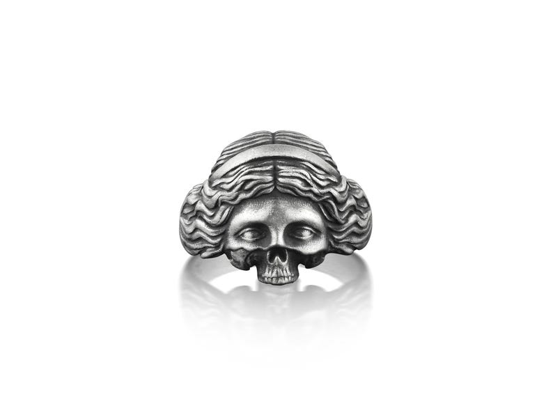 May include: A silver ring with a detailed skull design, featuring a woman's face with long, flowing hair.