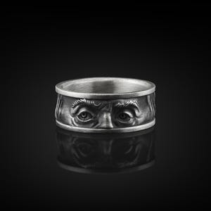 Könnte beinhalten: Ein silberner Ring mit einem detaillierten Design eines Gesichts mit zwei Augen. Der Ring befindet sich auf einem schwarzen Hintergrund.