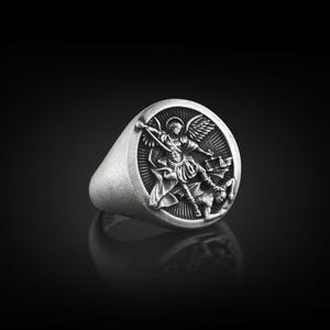 May include: A silver ring featuring a detailed design of an angel overcoming a demon. The ring is oval shaped and has a raised design.