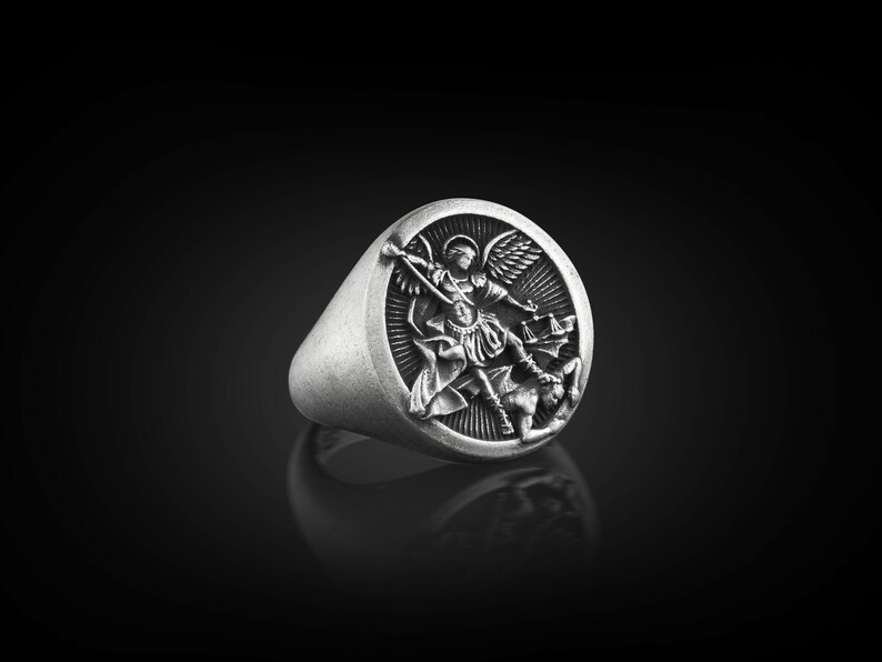 May include: A silver ring featuring a detailed design of an angel overcoming a demon. The ring is oval shaped and has a raised design.