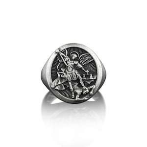 May include: A silver ring featuring a raised design of an angel overcoming a demon. The angel is holding a sword and the demon is lying on the ground.