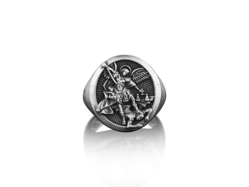 May include: A silver ring featuring a raised design of an angel overcoming a demon. The angel is holding a sword and the demon is lying on the ground.