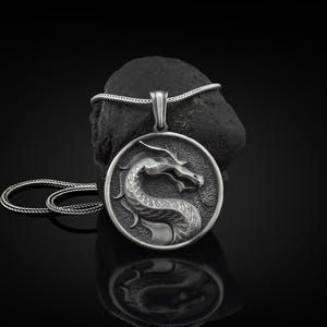 May include: Silver pendant necklace featuring a detailed dragon design. The pendant is round and has a textured surface. The necklace is on a black background.
