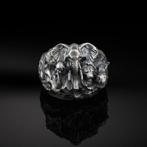 Può includere: Un anello d'argento con un design dettagliato di un elefante, un leone e altri animali. L'anello è un pezzo di dichiarazione con una superficie testurizzata.