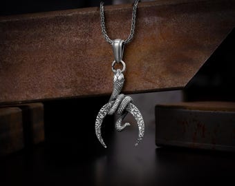 May include: A silver-toned pendant necklace featuring a detailed snake coiled around a crescent-shaped design. The pendant has intricate patterns and hangs from a matching chain. The background is a dark, textured surface.