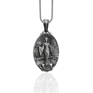 Puede incluir: Collar con colgante de plata con un dije ovalado detallado con una imagen en relieve de una mujer con los brazos extendidos. El dije tiene una superficie texturizada y un halo alrededor de la cabeza de la mujer.