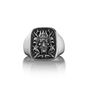 May include: A silver ring with a square face featuring a detailed, engraved design of a samurai warrior's mask.