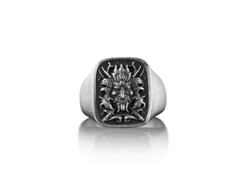 May include: A silver ring with a square face featuring a detailed, engraved design of a samurai warrior's mask.