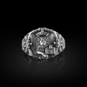 May include: A silver ring with a detailed design featuring a winged lion holding a book with the text "PAX TIBI MARCE EVANGELISTA MEUS", a floral design, and the words "ST. MARK".