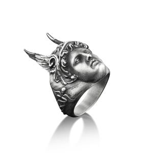 May include: A silver ring with a detailed sculpted face of a winged figure wearing a crown. The ring is angled to show the side profile of the face.