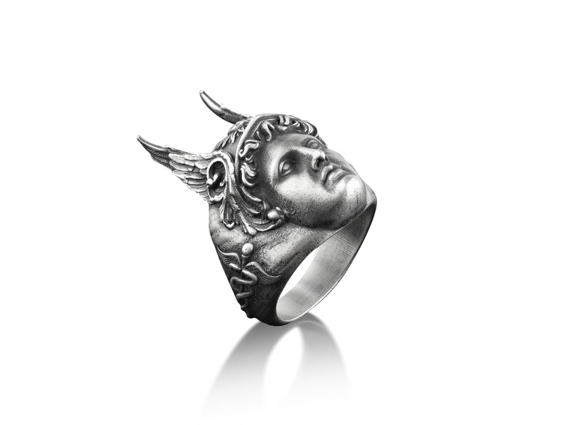 May include: A silver ring with a detailed sculpted face of a winged figure wearing a crown. The ring is angled to show the side profile of the face.