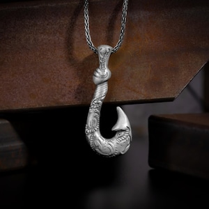 May include: A silver pendant necklace with a detailed fish hook design. The hook is intricately carved with floral and geometric patterns. The pendant hangs from a braided chain, creating a striking accessory.