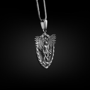 Puede incluir: Un collar con un colgante de plata que presenta un dise&ntilde;o esculpido detallado de un &aacute;ngel con alas sosteniendo una espada, sobre un fondo en forma de escudo.