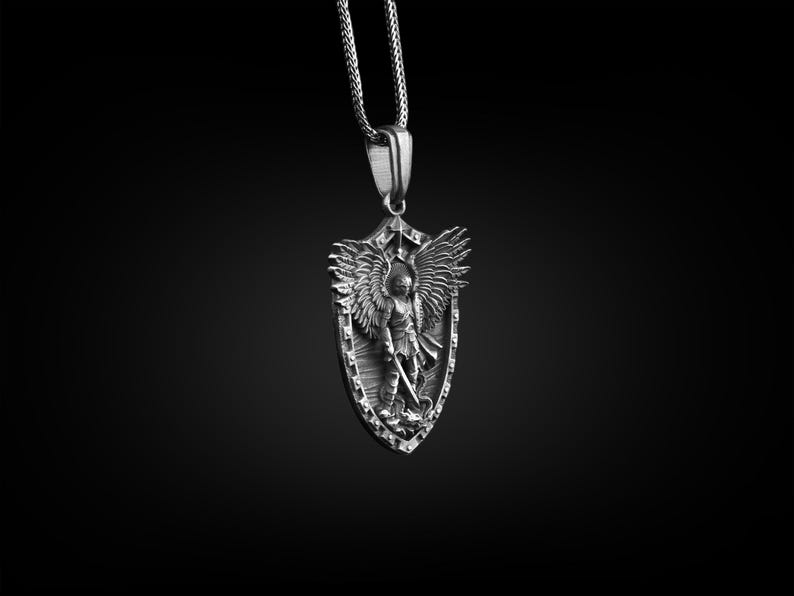 Puede incluir: Un collar con un colgante de plata que presenta un dise&ntilde;o esculpido detallado de un &aacute;ngel con alas sosteniendo una espada, sobre un fondo en forma de escudo.