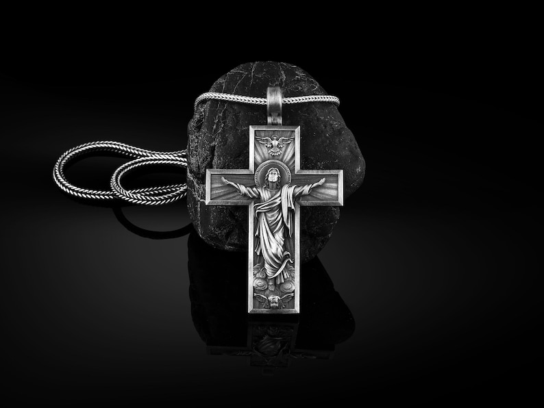 May include: A silver pendant necklace with a detailed cross featuring a depiction of Jesus Christ. The cross is on a black rock and the necklace is made of a braided silver chain.
