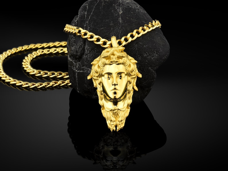 May include: A gold chain necklace with a pendant featuring a detailed, stylized face of a woman. The pendant is a three-dimensional design with a textured surface.