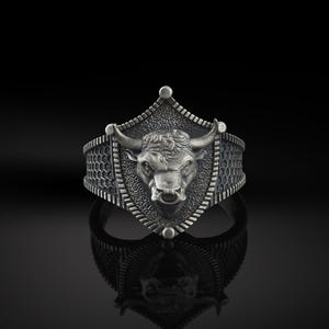 May include: A silver ring featuring a bull's head design on a shield-shaped setting. The ring has a textured band.