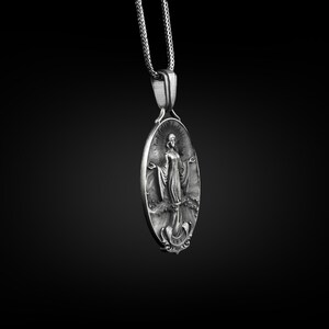 Puede incluir: Collar con colgante de plata con un dije ovalado detallado con una figura religiosa. El dije tiene un dise&ntilde;o en relieve y una superficie texturizada.