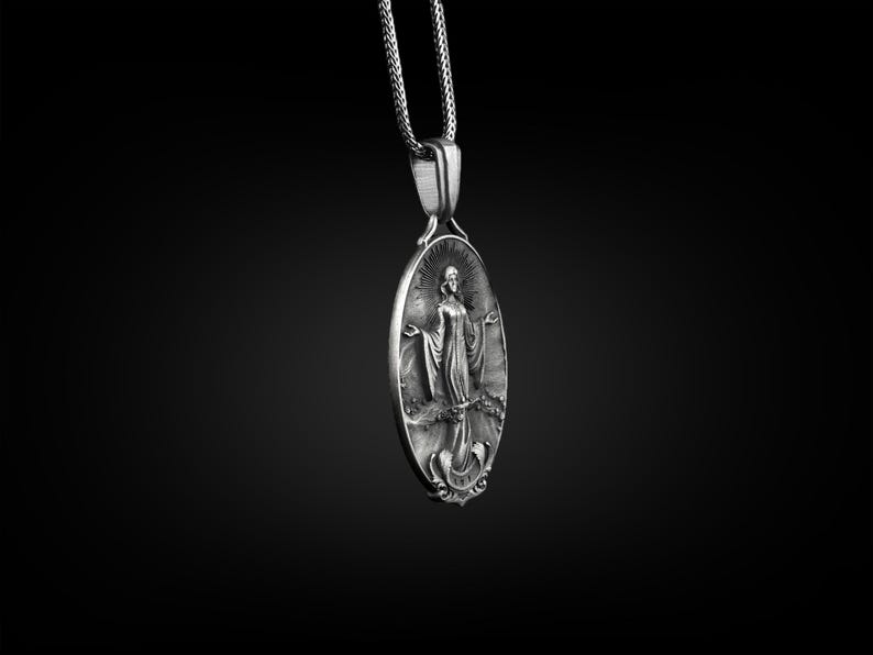 Puede incluir: Collar con colgante de plata con un dije ovalado detallado con una figura religiosa. El dije tiene un dise&ntilde;o en relieve y una superficie texturizada.