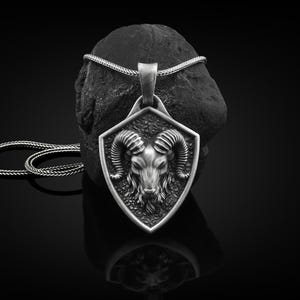 May include: A silver-colored pendant necklace featuring a ram's head within a shield-shaped frame. The pendant hangs from a silver chain, displayed against a dark, textured background. The ram's head has detailed horns and facial features.