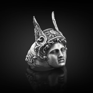 May include: A silver ring with a detailed design of a woman's face wearing a winged helmet with a sun symbol. The ring is a statement piece with intricate details.