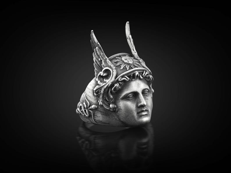 May include: A silver ring with a detailed design of a woman's face wearing a winged helmet with a sun symbol. The ring is a statement piece with intricate details.