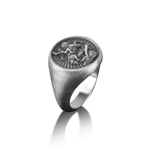 Puede incluir: Un anillo de plata con un dise&ntilde;o ovalado en relieve que presenta una representaci&oacute;n detallada de una figura alada. El anillo tiene un acabado cepillado.