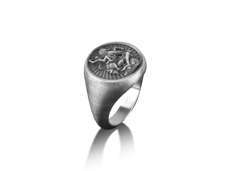 Puede incluir: Un anillo de plata con un dise&ntilde;o ovalado en relieve que presenta una representaci&oacute;n detallada de una figura alada. El anillo tiene un acabado cepillado.